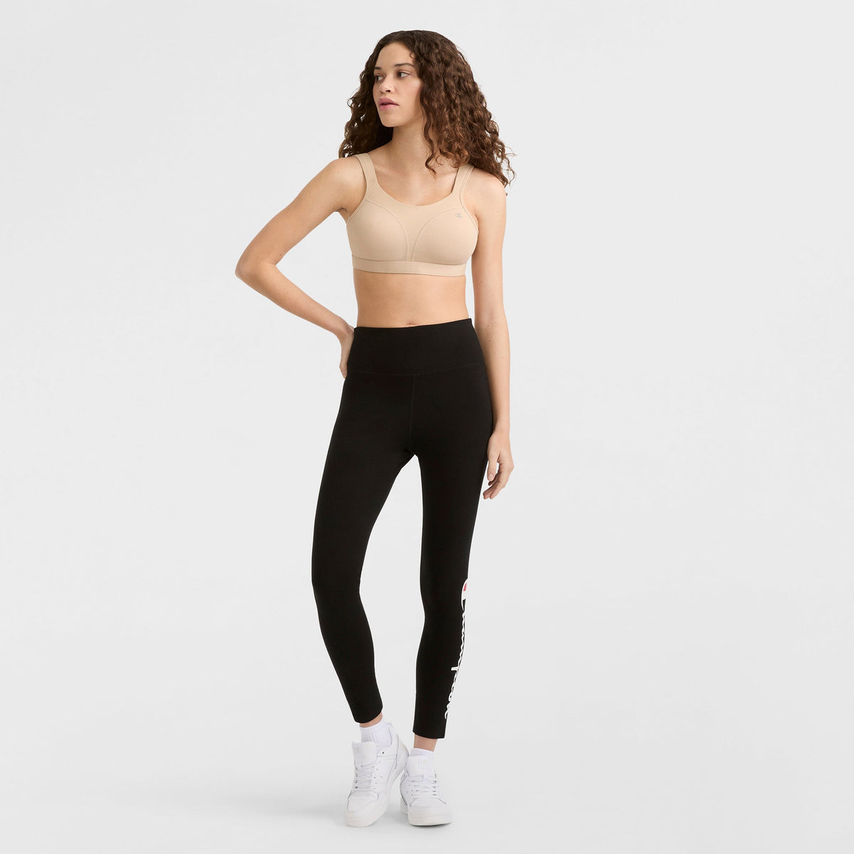 A woman with long curly hair wears the Champion Spot Comfort High-Impact Sports Bra, C Logo in beige, high-waisted black leggings, and white sneakers. She poses against a plain white background with one hand on her hip.