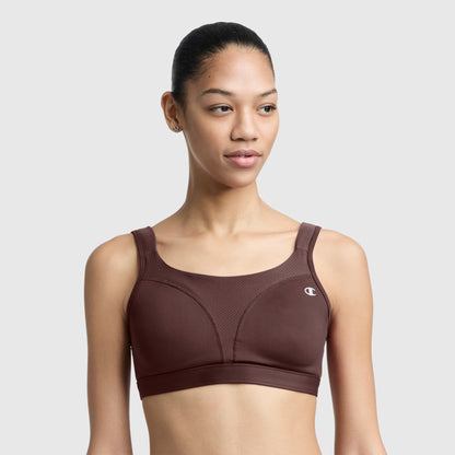 A woman with tied-back hair wears the Champion Spot Comfort High-Impact Sports Bra featuring the C Logo in brown, standing against a plain light gray background and looking slightly to the side.