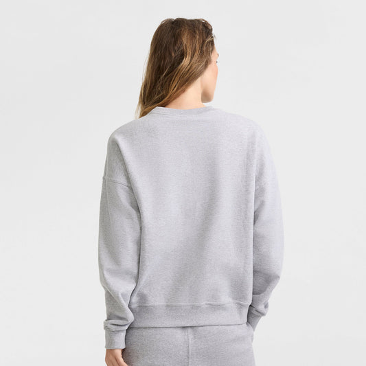 A person with long hair faces away, wearing a Champion Powerblend Crewneck Sweatshirt with script logo in light gray and matching sweatpants against a plain white background.