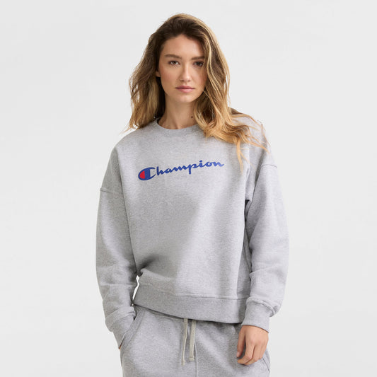 A woman with wavy blonde hair wears a light gray Champion Powerblend Crewneck Sweatshirt with Script Logo and matching sweatpants, standing against a plain white background.