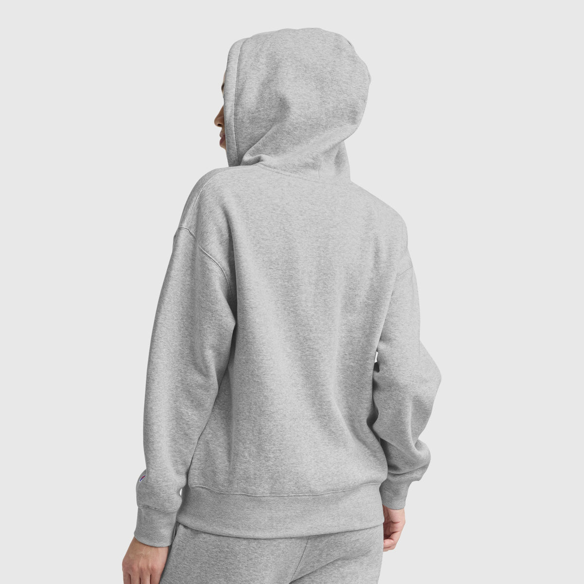 A person wearing a light gray Champion Powerblend Fleece Zip-Up Hoodie, C Logo, with the hood up, and matching sweatpants is seen from the back against a plain light background.