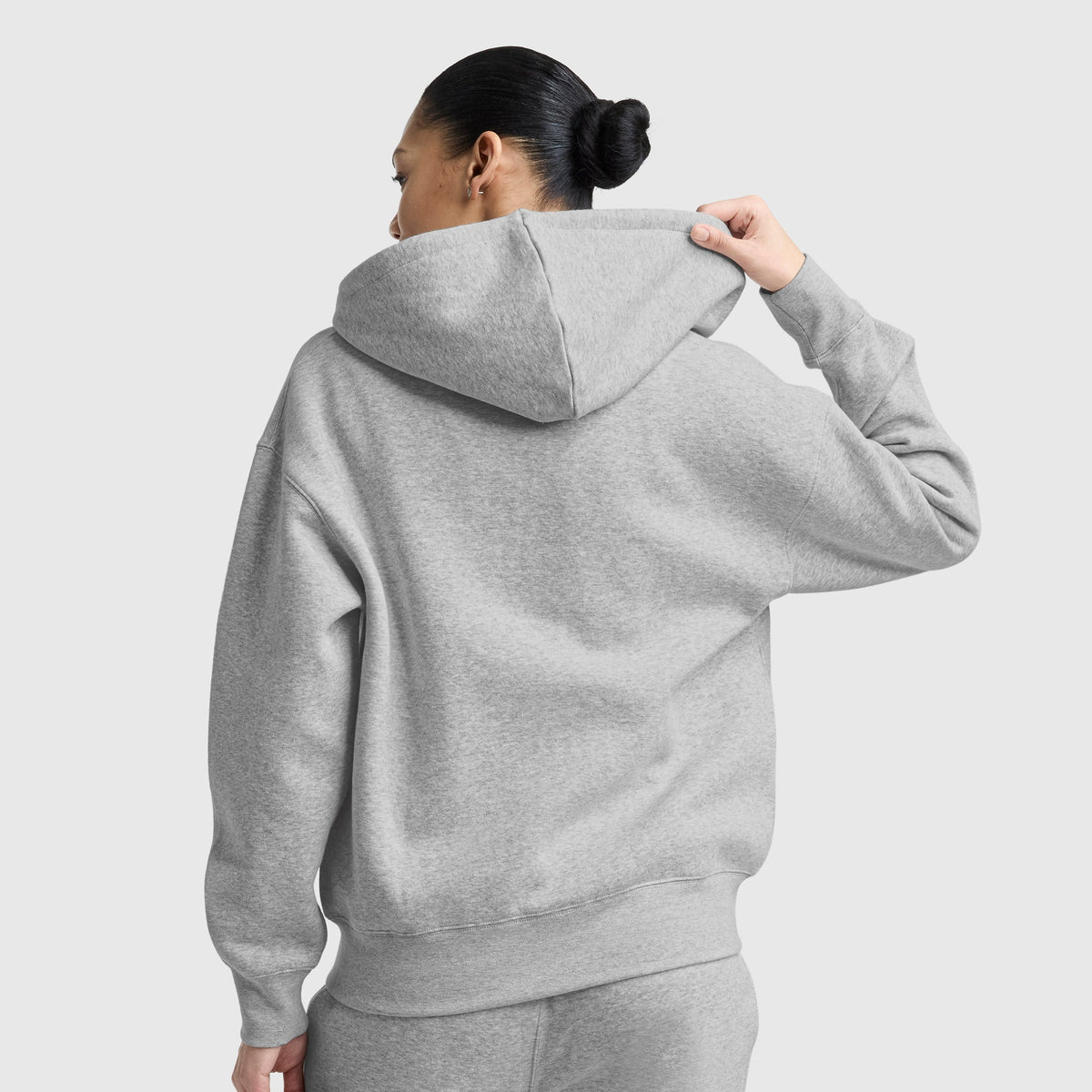 A person with dark hair in a bun wears the Champion Powerblend Fleece Zip-Up Hoodie, C Logo, in gray and sweatpants, facing away and lifting the hood against a plain light gray background.