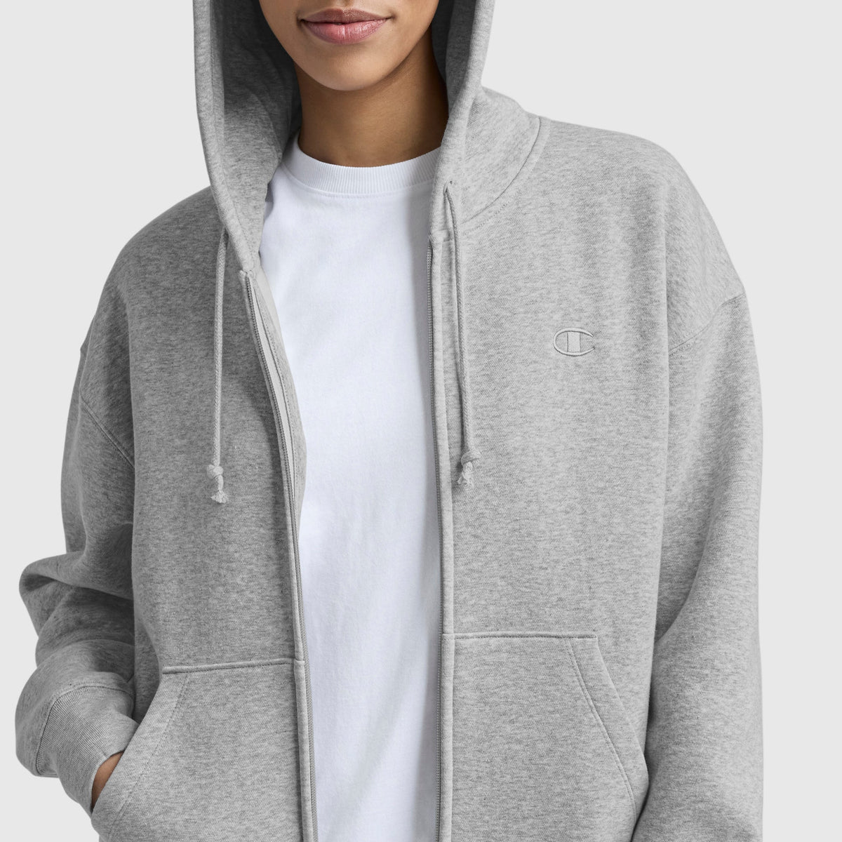 A person in a light gray Champion Powerblend Fleece Zip-Up Hoodie, C Logo, with the hood up over a plain white T-shirt and hands in the pockets. Only the lower part of their face is visible.
