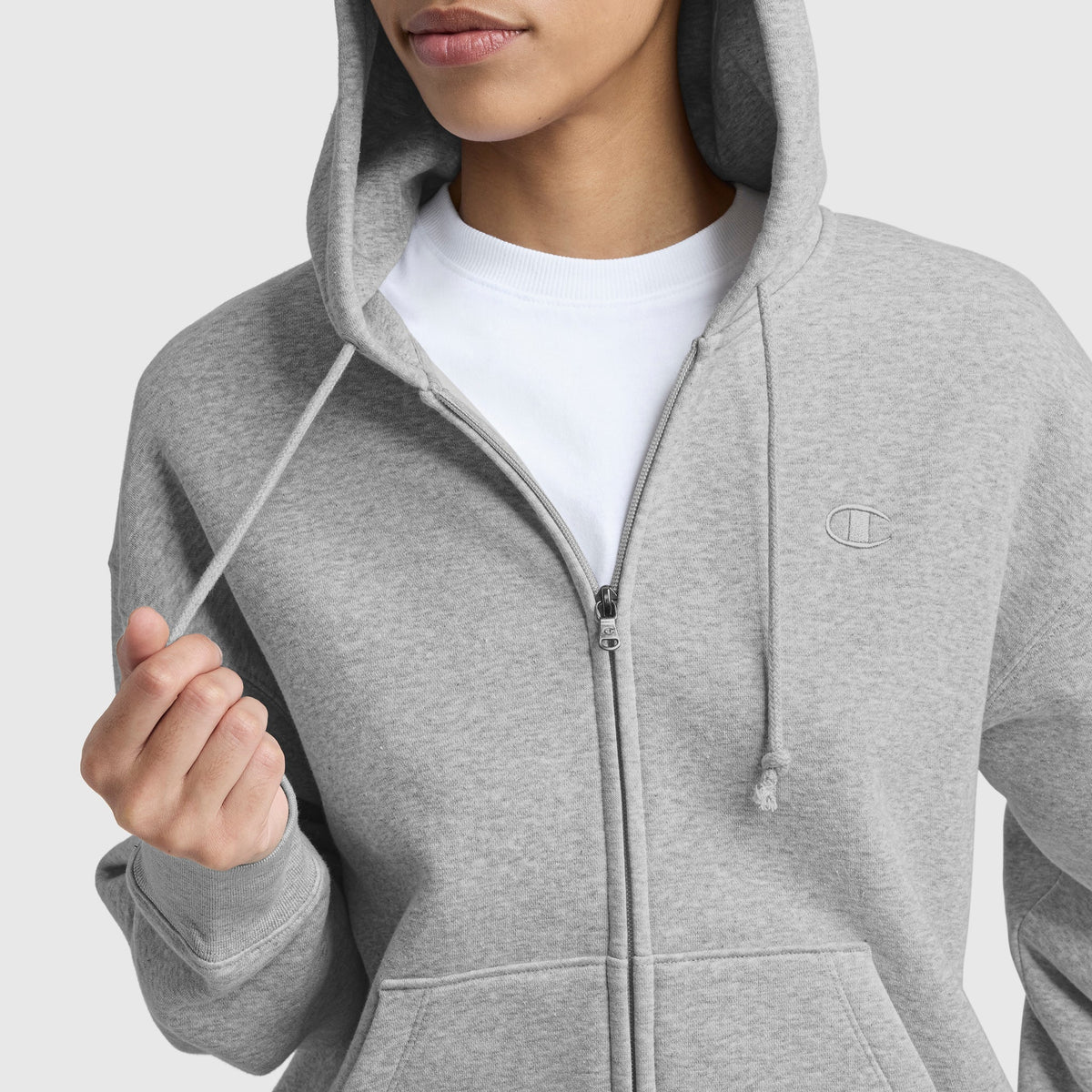 A person wears a light gray Champion Powerblend Fleece Zip-Up Hoodie, C Logo, over a white t-shirt, holding one drawstring. The face is partially visible and the hoodie features a small embroidered logo on the chest.