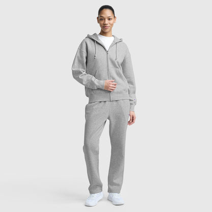 A person stands against a plain background, smiling softly with one hand on their jacket. They're wearing a Champion Powerblend Fleece Zip-Up Hoodie with C Logo in light gray, matching sweatpants, a white t-shirt, and white sneakers.