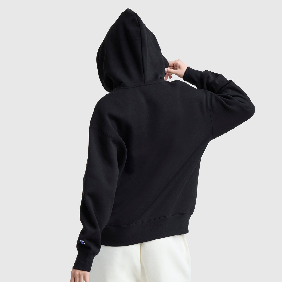 A person in a black Champion Powerblend Fleece Zip-Up Hoodie, C Logo, and white pants stands facing away, touching the hood against a plain light gray background.