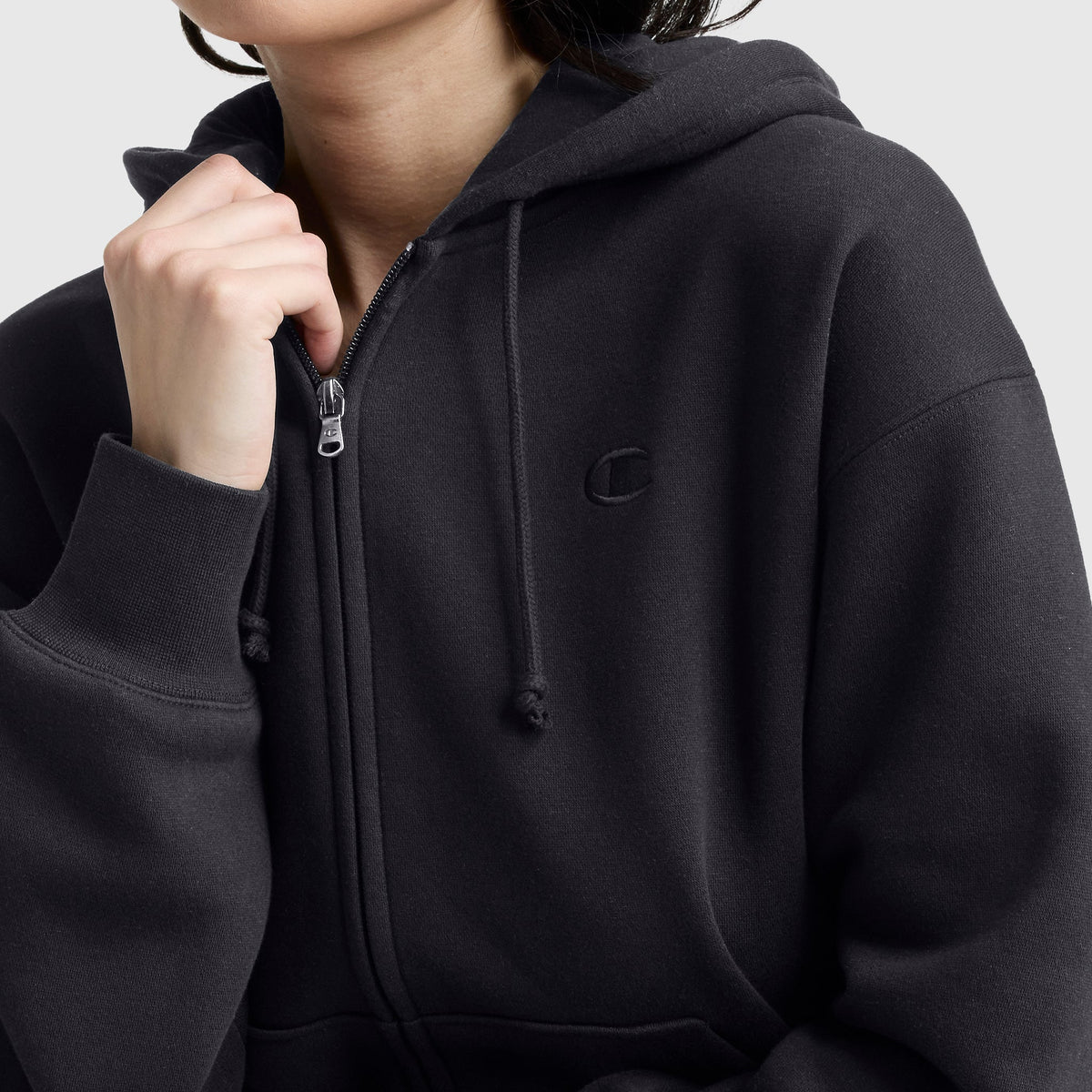 A person wears a dark gray Champion Powerblend Fleece Zip-Up Hoodie with a hood, drawstrings, and the C logo stitched on the chest, holding the zipper near their chin. The background is plain and light-colored.