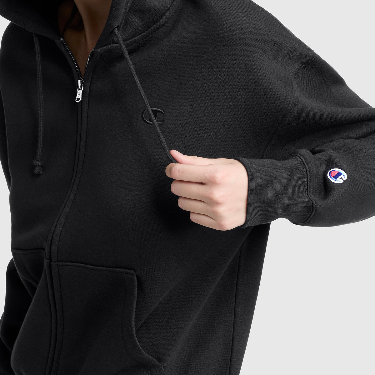 A person wears a black Champion Powerblend Fleece Zip-Up Hoodie, C Logo, pulling the drawstring with one hand. Champion branding is visible on the chest and sleeve against a plain gray background.