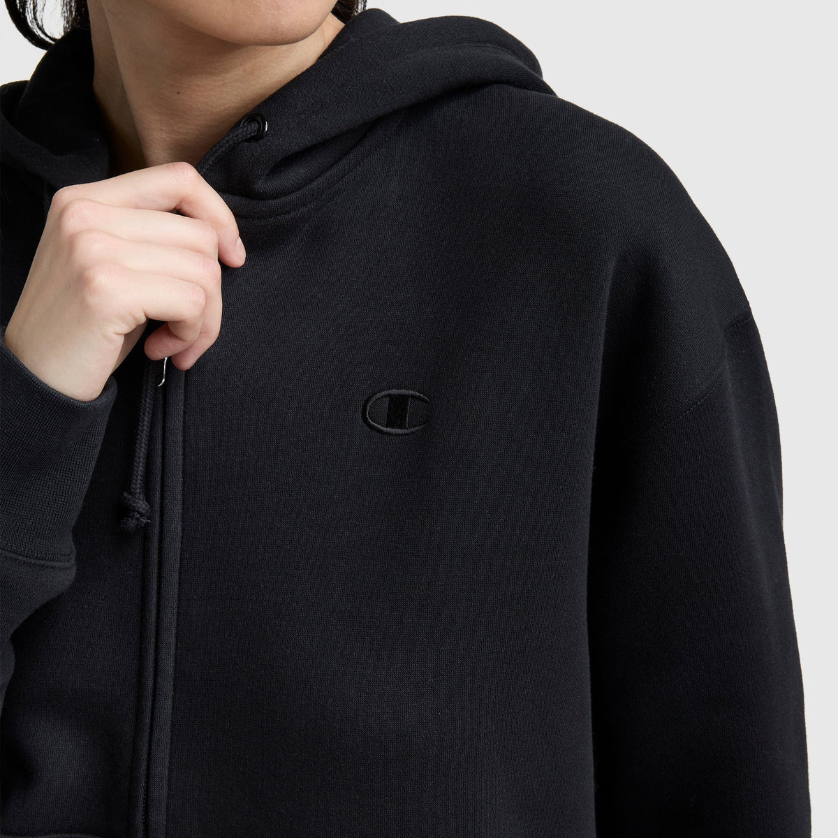 A person in a black Champion Powerblend Fleece Zip-Up Hoodie, C Logo, adjusts the drawstring. The embroidered Champion logo is visible on the chest, with their face partially out of view against a plain light background.