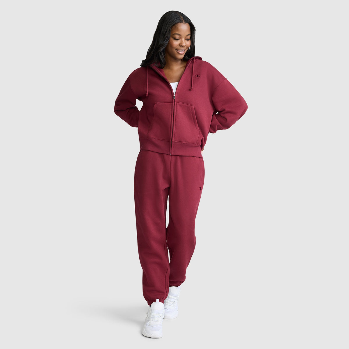 A woman stands smiling, wearing a matching maroon Champion Powerblend Fleece Zip-Up Hoodie with the C Logo and sweatpants set paired with white sneakers against a plain light gray background.
