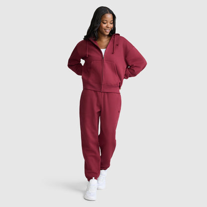 A woman stands smiling, wearing a matching maroon Champion Powerblend Fleece Zip-Up Hoodie with the C Logo and sweatpants set paired with white sneakers against a plain light gray background.