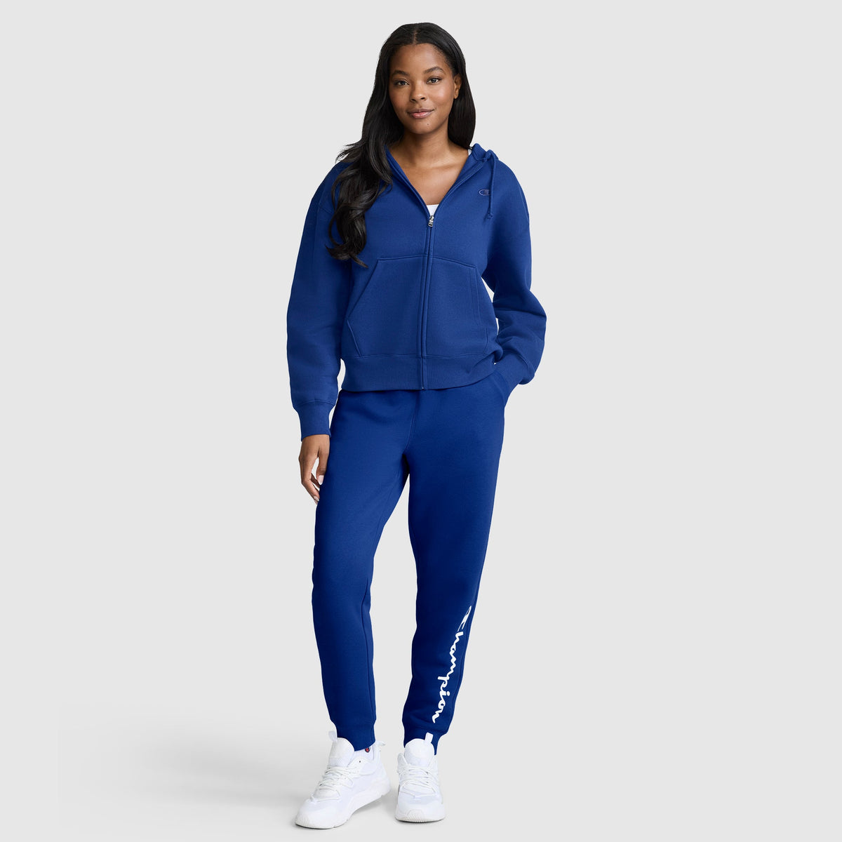 A woman wearing a matching blue Champion Powerblend Fleece Zip-Up Hoodie, C Logo, and sweatpants with white sneakers stands against a plain light gray background, looking at the camera with a relaxed expression and one hand in her pocket.