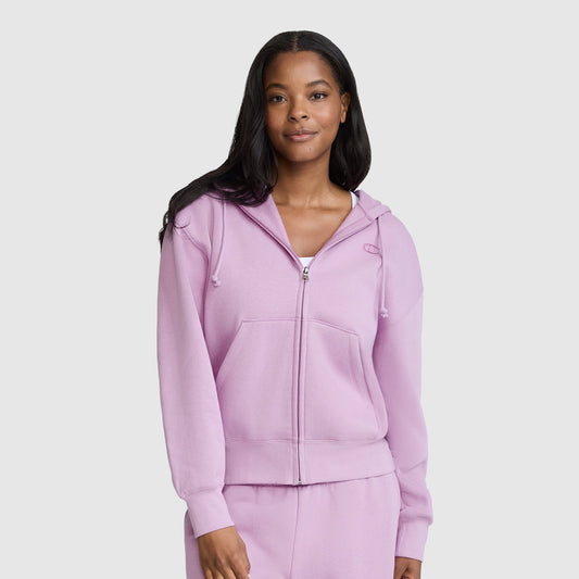 A woman stands against a plain light gray background, smiling softly with her hands by her sides, wearing a Champion Powerblend Fleece Zip-Up Hoodie, C Logo in light purple along with matching sweatpants.