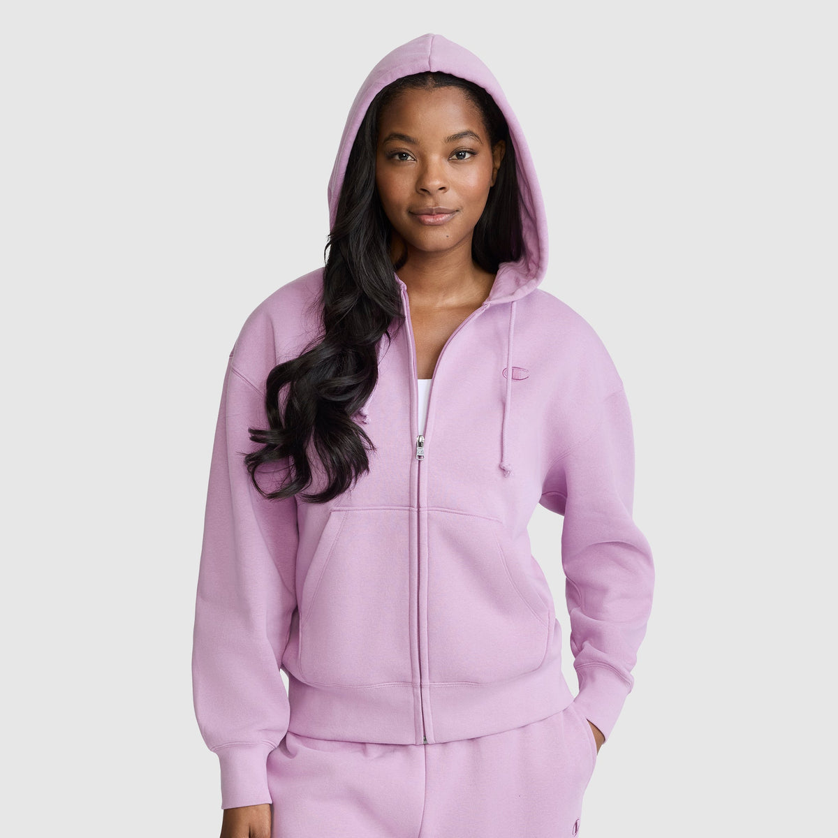 A woman stands facing forward with her hands in the pockets of a light purple Champion Powerblend Fleece Zip-Up Hoodie, featuring a front zipper and a hood pulled over her head. She pairs the hoodie with matching sweatpants.