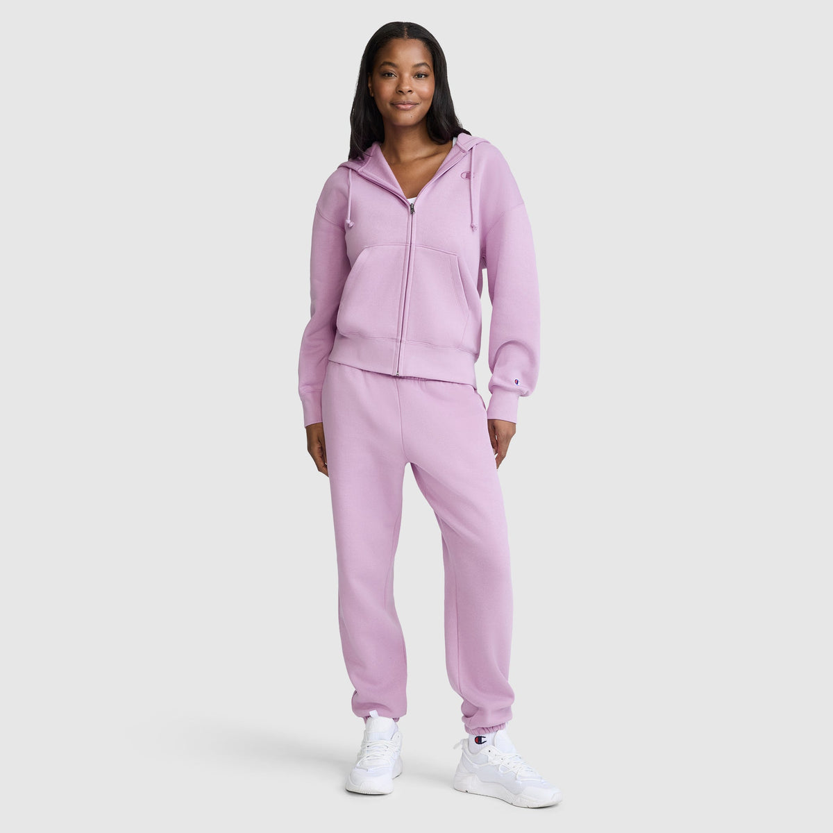 A woman stands smiling, wearing a Champion Powerblend Fleece Zip-Up Hoodie with the C logo in light purple, paired with matching sweatpants and white sneakers against a plain light gray background.
