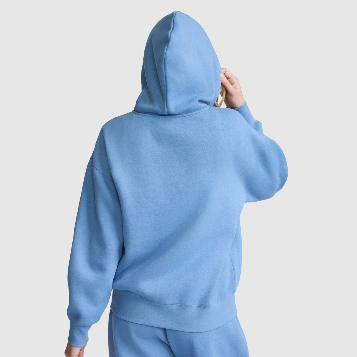 A person wearing a loose-fitting, light blue Champion Powerblend Fleece Zip-Up Hoodie, C Logo, along with matching pants is facing away from the camera with the hood up and one hand touching the side of the hood. The background is plain white.