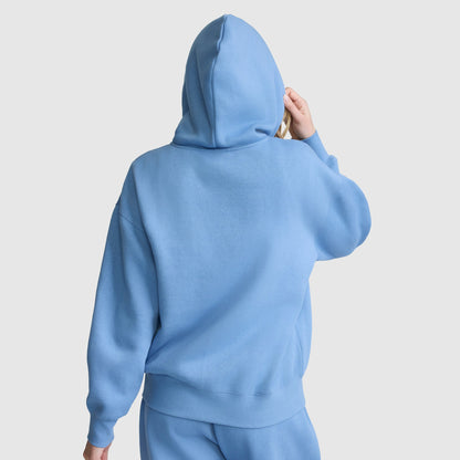 A person wearing a loose-fitting, light blue Champion Powerblend Fleece Zip-Up Hoodie, C Logo, along with matching pants is facing away from the camera with the hood up and one hand touching the side of the hood. The background is plain white.