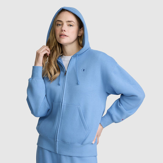 A woman wearing the Champion Powerblend Fleece Zip-Up Hoodie, C Logo, with the hood up and paired with matching sweatpants, stands against a plain light gray background.