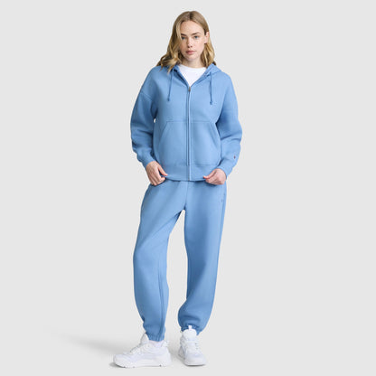 A woman stands against a plain light background, wearing a light blue Champion Powerblend Fleece Zip-Up Hoodie with the C Logo, paired with matching sweatpants, white sneakers, and a white t-shirt.
