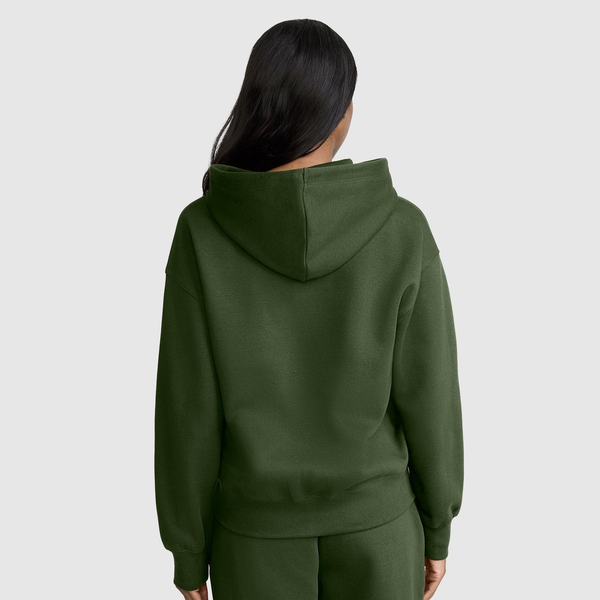 A person with long dark hair is wearing a Champion Powerblend Fleece Zip-Up Hoodie, C Logo, in dark green along with matching pants, facing away from the camera against a plain light background.