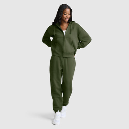 A woman wearing a Champion Powerblend Fleece Zip-Up Hoodie, C Logo, in olive green and matching sweatpants with white sneakers stands against a plain light gray background, smiling and looking slightly down.