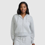 A woman stands facing forward against a plain light background, hands in the front pockets of her light gray Champion Cropped Zip-Up Hoodie paired with matching sweatpants.
