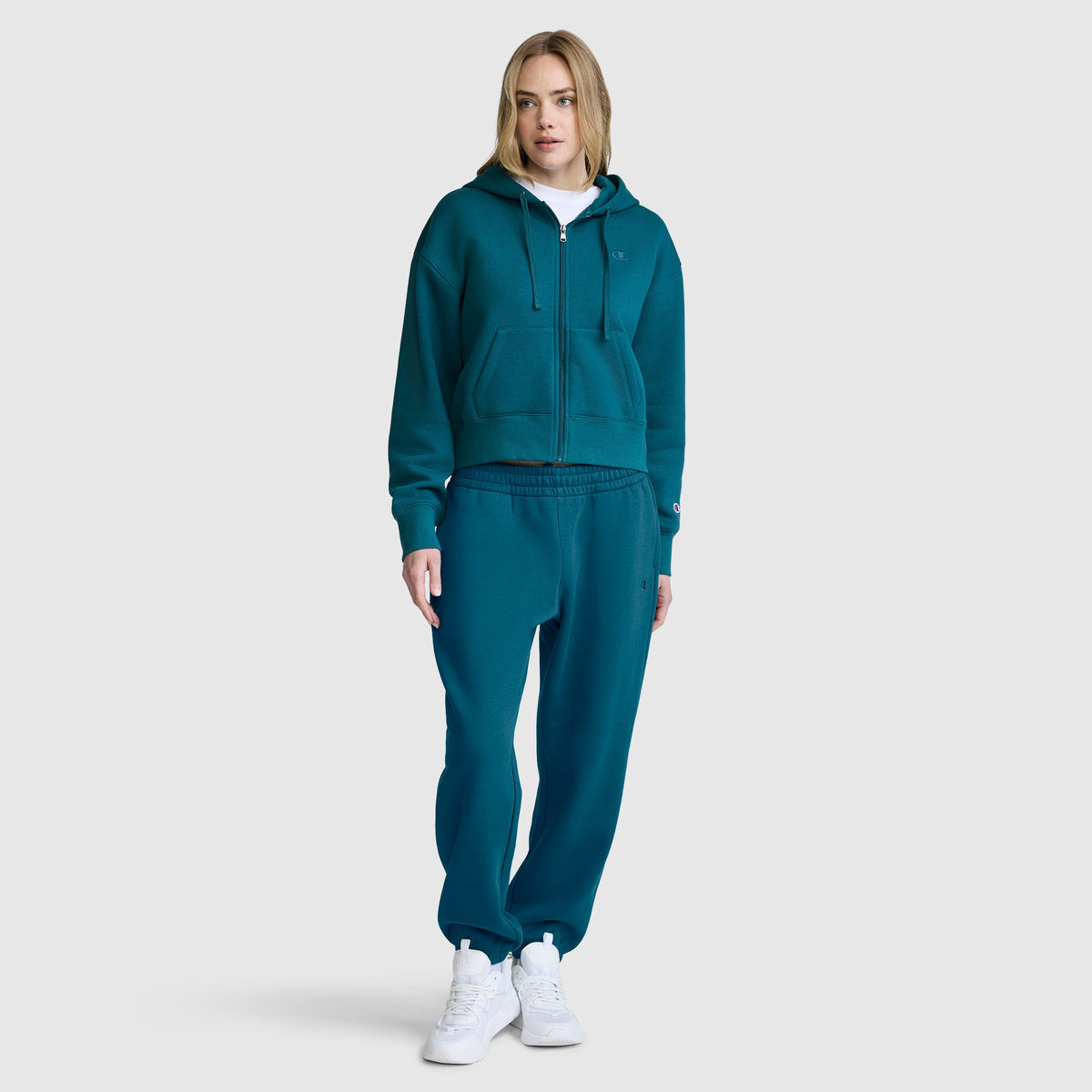 A woman stands facing forward against a plain light gray background, wearing a teal Champion Cropped Zip-Up Hoodie with matching sweatpants and white sneakers.