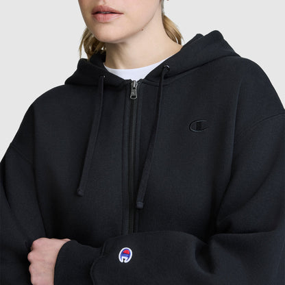 A person wearing a black Champion Cropped Zip-Up Hoodie with a small embroidered logo on the chest and sleeve stands against a plain light background. This cozy fleece hoodie combines comfort and style with its zip-up design.