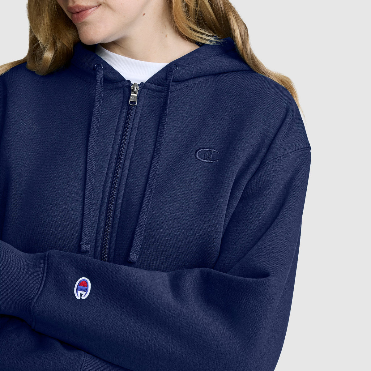 A person wearing a navy blue Champion Cropped Zip-Up Hoodie, featuring the iconic logo on the sleeve and chest, stands against a plain light background.