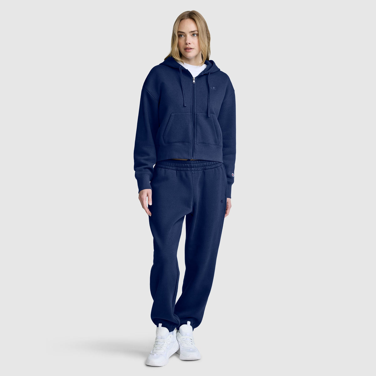 A woman stands facing forward against a plain light gray background, wearing a navy blue Champion Cropped Zip-Up Hoodie, matching sweatpants, a white t-shirt, and white sneakers.