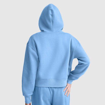 A person is seen from behind, wearing a plain, light blue Champion Cropped Zip-Up Hoodie and matching sweatpants against a neutral background. Their right hand rests on their hip.
