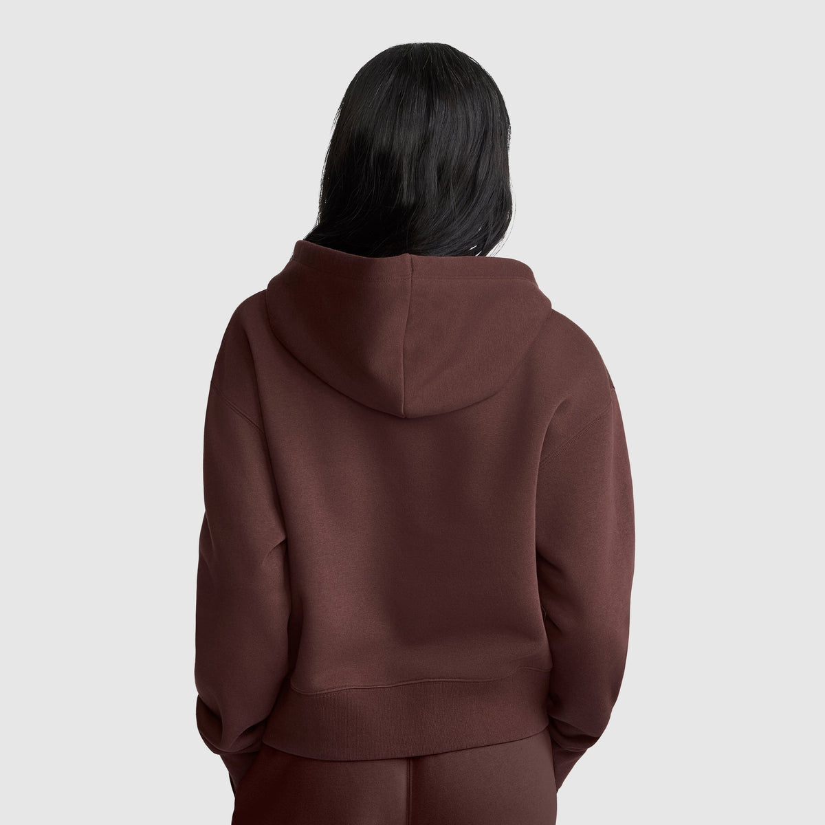 A person with long dark hair is wearing a brown Champion Cropped Zip-Up Hoodie and matching pants, standing with their back facing the camera against a plain light gray background.