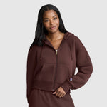 A woman wearing a brown Champion Cropped Zip-Up Hoodie and matching pants stands against a plain background, smiling slightly with one hand in her pocket and the other holding the fleece hoodie.