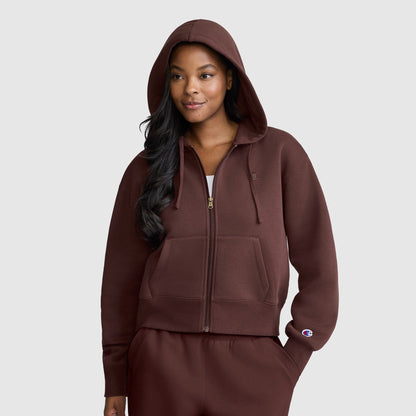 A woman wearing the Champion Cropped Zip-Up Hoodie in brown with the hood up, paired with matching sweatpants, stands against a plain light gray background.