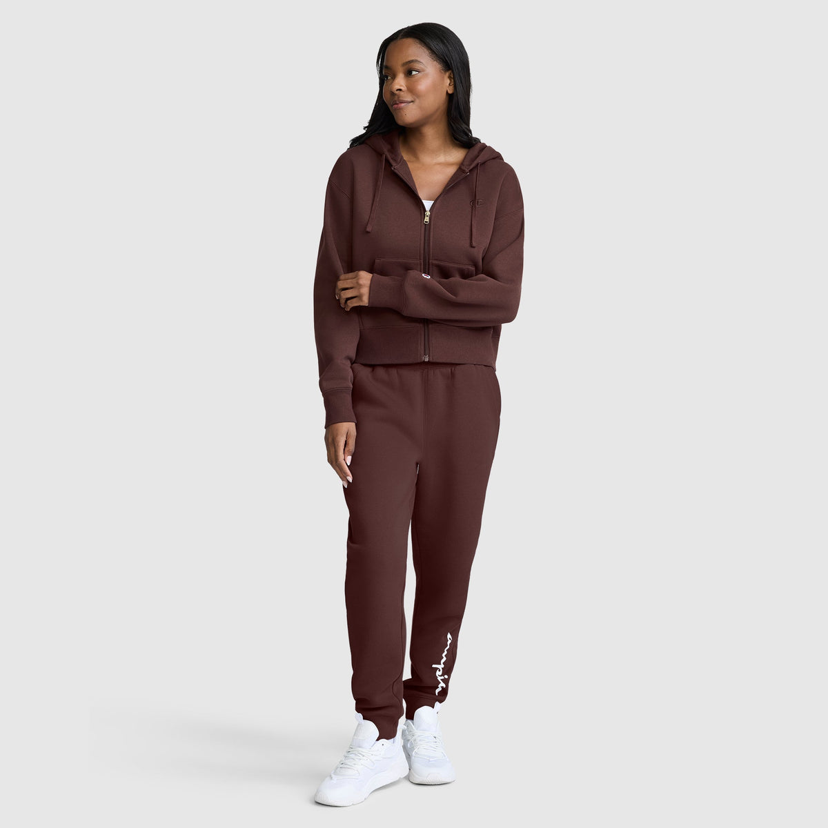 A woman stands wearing a matching maroon Champion Cropped Zip-Up Hoodie and joggers with white sneakers, looking to the side and smiling against a plain light gray background.