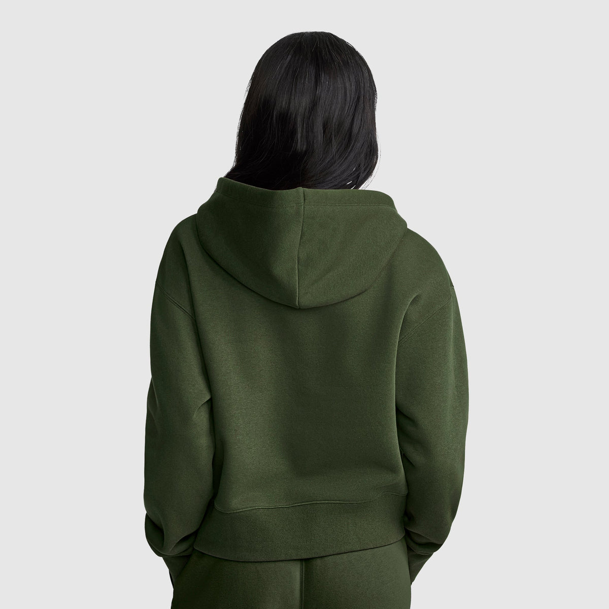 A person with long dark hair stands with their back to the camera against a plain light gray background, wearing the Champion Cropped Zip-Up Hoodie in olive green, paired with matching pants.