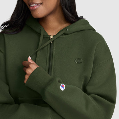 A woman wearing a Champion Cropped Zip-Up Hoodie in dark green fleece, featuring the Champion logo on the chest and sleeve, holds one of the hoodie’s drawstrings and smiles slightly.