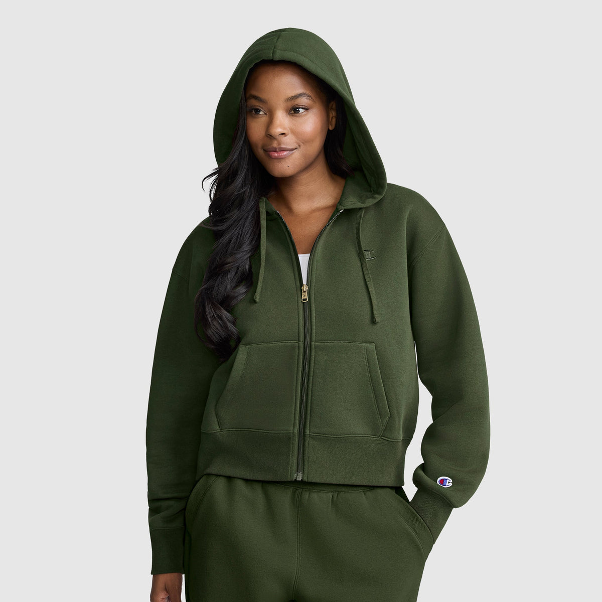 A woman wearing a dark green Champion Cropped Zip-Up Hoodie with the hood up and matching sweatpants stands facing forward, smiling slightly, against a plain light gray background.