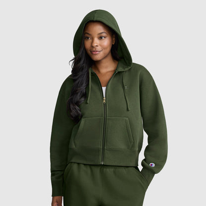 A woman wearing a dark green Champion Cropped Zip-Up Hoodie with the hood up and matching sweatpants stands facing forward, smiling slightly, against a plain light gray background.
