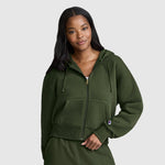 A woman with long dark hair wears a green Champion Cropped Zip-Up Hoodie and matching pants. She stands against a plain gray background, smiling softly with one hand in her pocket.