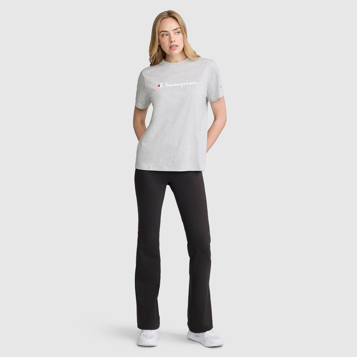 A woman stands facing forward against a plain light background, wearing a light gray Champion t-shirt and black Champion Soft Touch Flare Leggings with an anti-odor finish and C logo. She has her hands in her pockets, a neutral expression on her face, and completes the look with white sneakers.