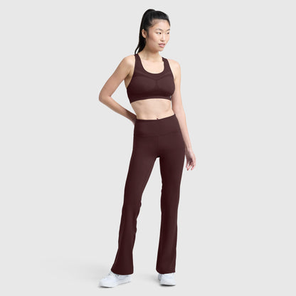 A woman stands against a plain background wearing Champion’s Soft Touch Flare Leggings with a C Logo in burgundy, paired with a matching sports bra and white sneakers. Her hair is pulled back in a ponytail as she looks to the side, highlighting these anti-odor, stylish women’s workout leggings.