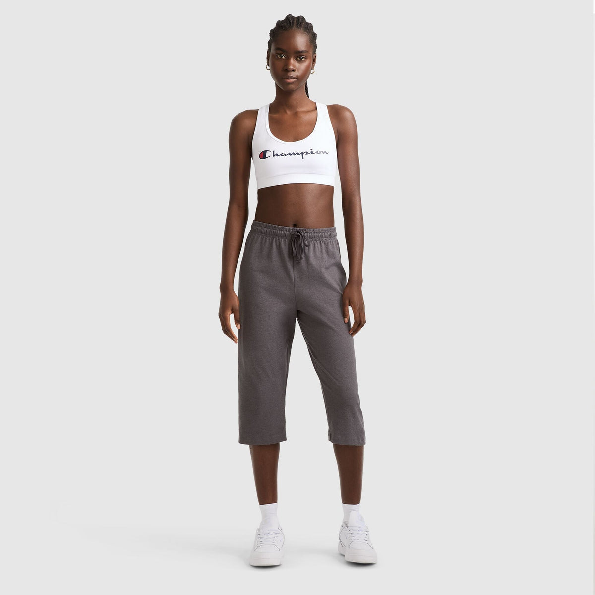 A woman stands on a plain background wearing a white Champion sports bra, Lightweight Lounge Capri Pants, C Logo in gray by Champion, plus white socks and sneakers. She faces forward with her arms at her sides.