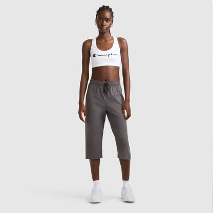 A woman stands on a plain background wearing a white Champion sports bra, Lightweight Lounge Capri Pants, C Logo in gray by Champion, plus white socks and sneakers. She faces forward with her arms at her sides.