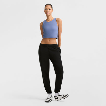 A woman stands against a plain white background, wearing Champion's Powerblend Oversized Sweatpants with a C Logo in black, paired with a sleeveless blue crop top and white sneakers. She has her hands behind her back and looks forward neutrally.