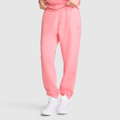 A person wearing light pink Champion Powerblend Oversized Sweatpants with the C Logo and white sneakers stands against a plain grey background. Only the lower half of the body is visible, with hands adjusting the waistband of the fleece pants.