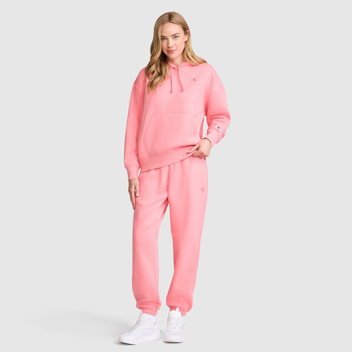 A woman wearing a matching light pink hoodie and Champion Powerblend Oversized Sweatpants with C Logo, paired with white sneakers, stands on a neutral background and smiles at the camera.