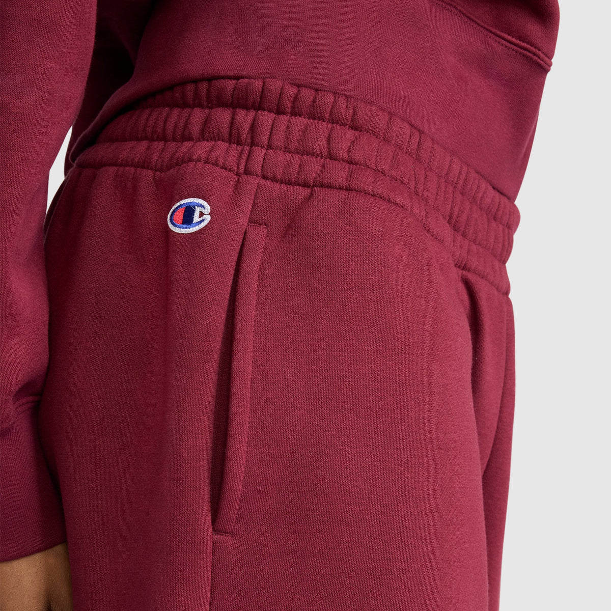 A woman is wearing Champion Powerblend Oversized Sweatpants with the C Logo in maroon, made from cozy fleece and designed with an elastic waistband and signature logo on the left pocket, paired with a matching maroon sweatshirt. The background is plain light gray.