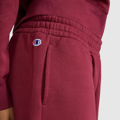 A woman is wearing Champion Powerblend Oversized Sweatpants with the C Logo in maroon, made from cozy fleece and designed with an elastic waistband and signature logo on the left pocket, paired with a matching maroon sweatshirt. The background is plain light gray.