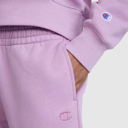 Wearing a matching lavender Champion sweatshirt and Champion Powerblend Oversized Sweatpants with the C Logo, the person stands with one hand in their pocket. Both garments showcase the iconic Champion branding and are made from soft Powerblend fleece for added comfort.